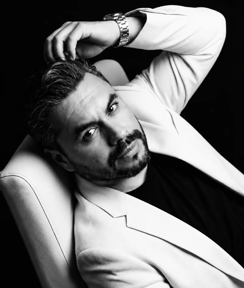 Retrato en blanco y negro de Karloz Vázquez con saco claro, recostado en un sillón